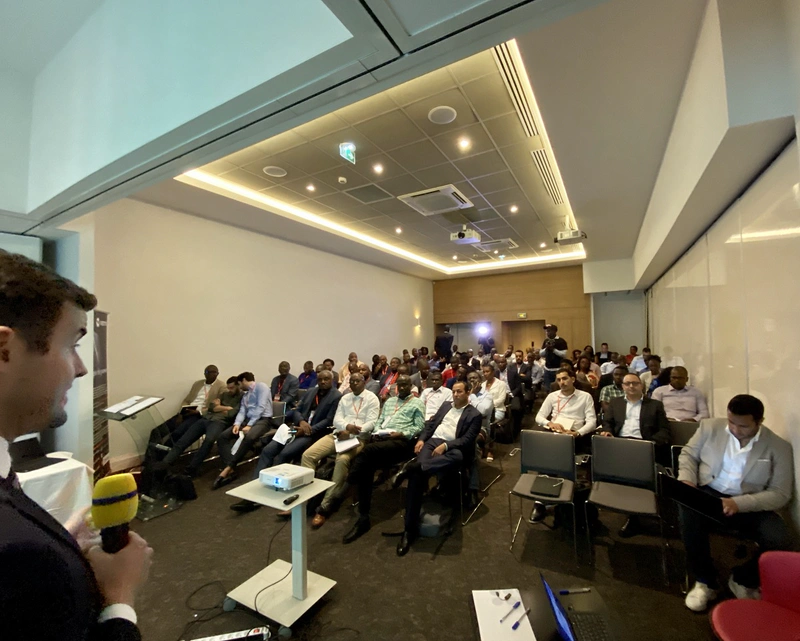 Seen from over his shoulder, a man talks into a mic and presents to a roomful of people 