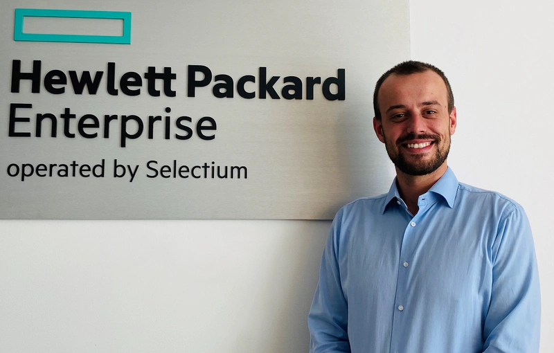 a man is seen in a blue shirt in a portrait photo, smiling in front of a wall with a Hewlett Packard Enterprise operated by Selectium logo 