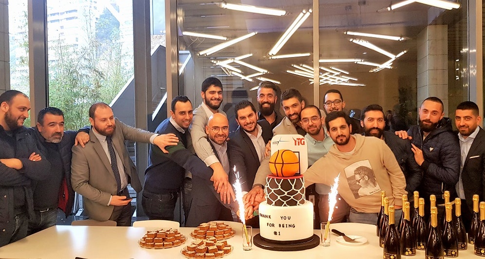 Group of men cluster together behind a celebration cake which has a basketball on top of it