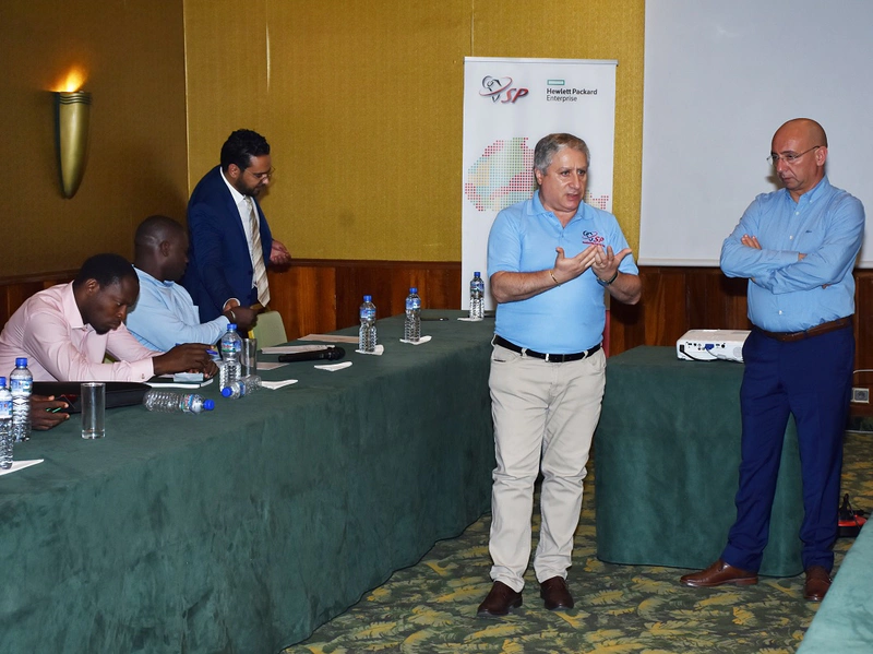 Two men in blue shirts stand near each other as one of them speaks to the other