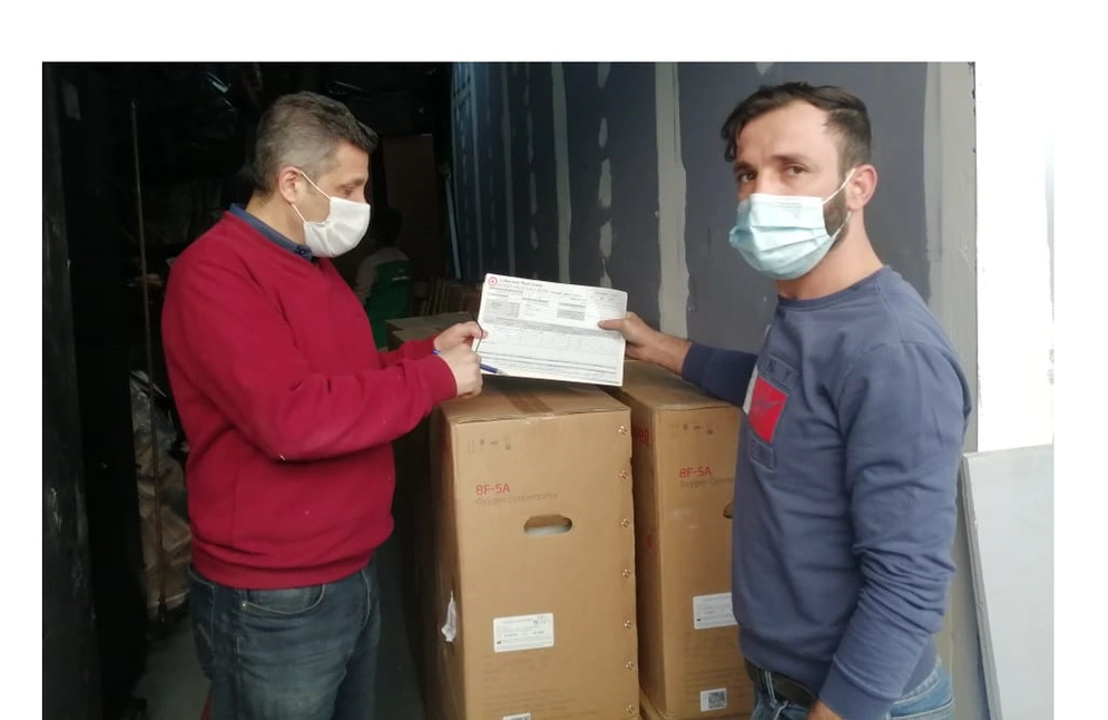 shows two men in covid masks examining a shipping consignment note in front of boxed machinery