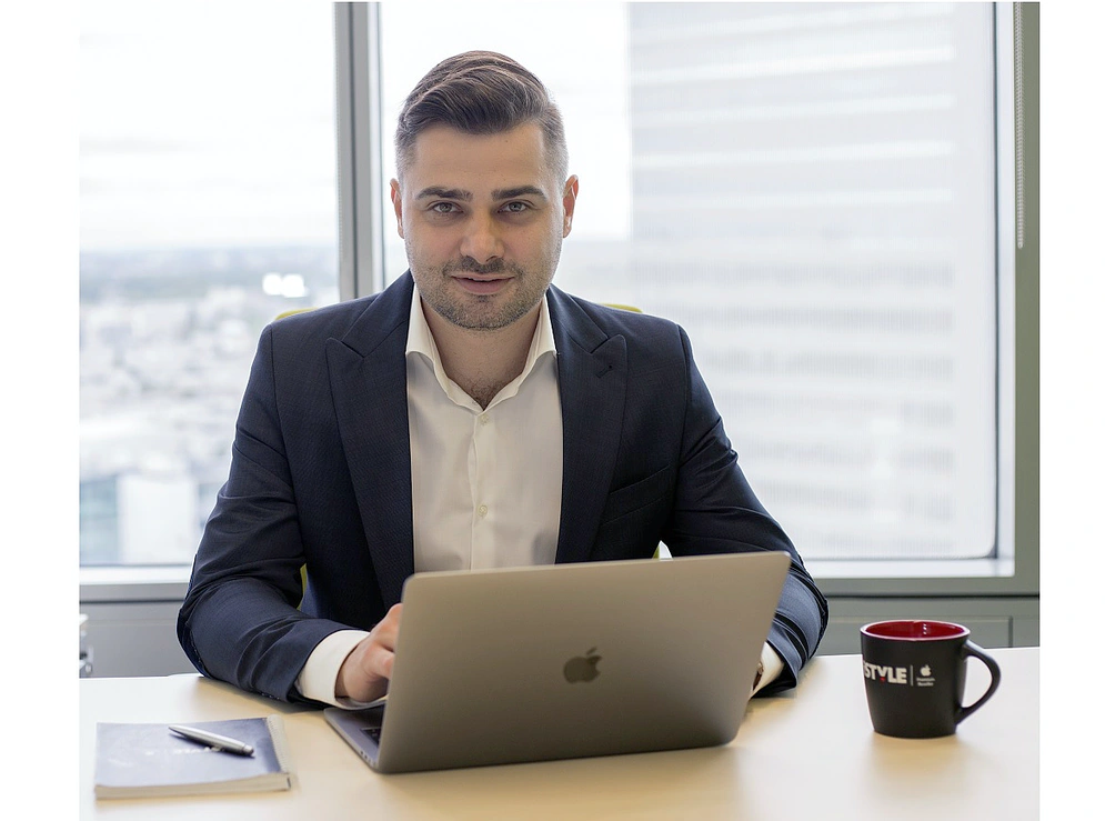 a man in a jacket and shirt sits at a desk behind a laptop screen