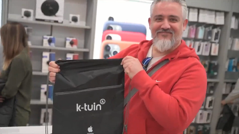 a man wearing a red hoodie holds a black carrier bag with the k-tuin logo and an Apple logo