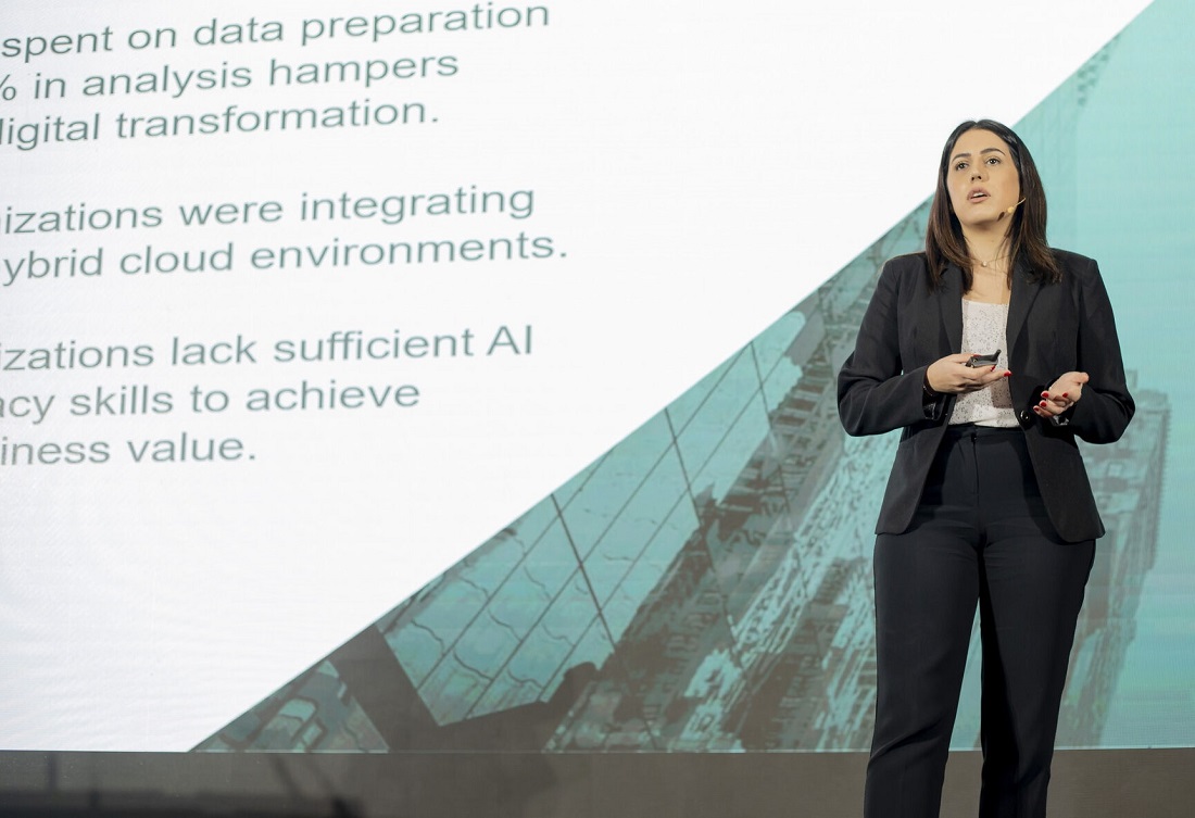 a woman in a business suit is presenting on stage and a powerpoint slide can be seen to the left 