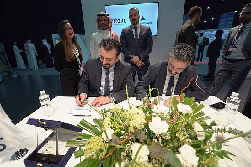 two men sit at a desk and sign a document with flowers in the foreground