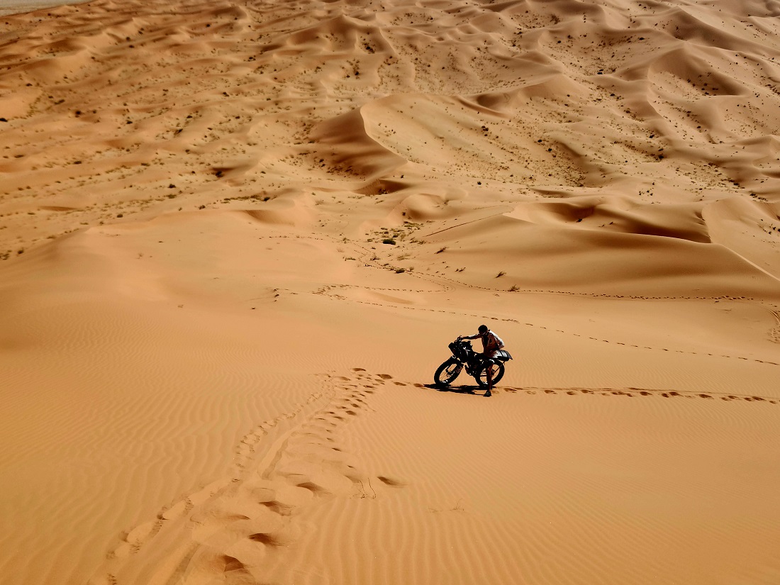 a lone cyclist is seen in a desert landscape with footprints visible in the foreground