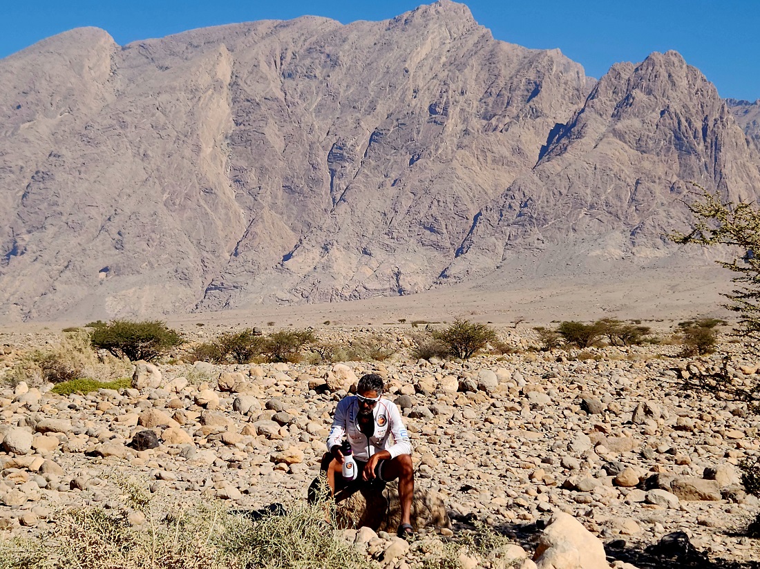 un homme est vu dans un paysage désertique, avec des montagnes en arrière-plan, se reposant sur un rocher et buvant à une bouteille d'eau. 