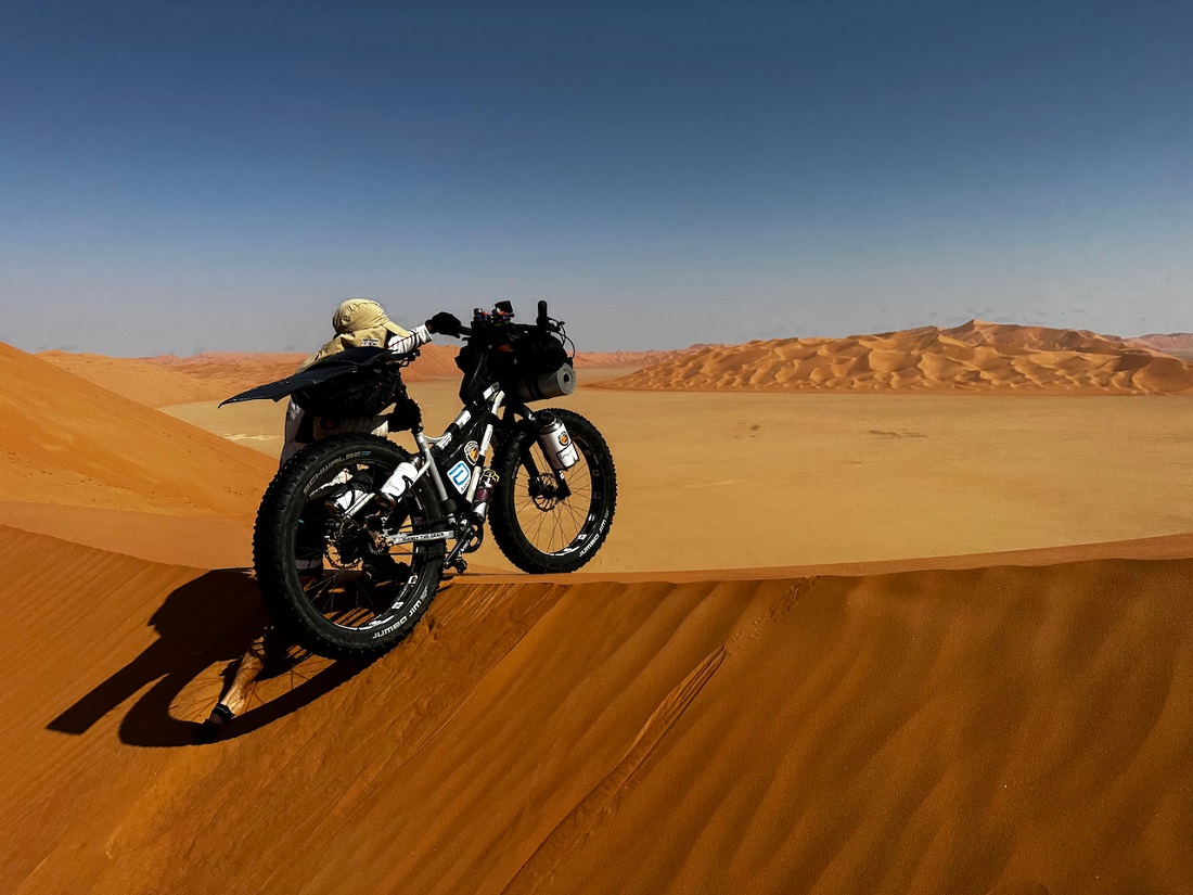 a man pushes a heavily laden bike up and over a high sand dune