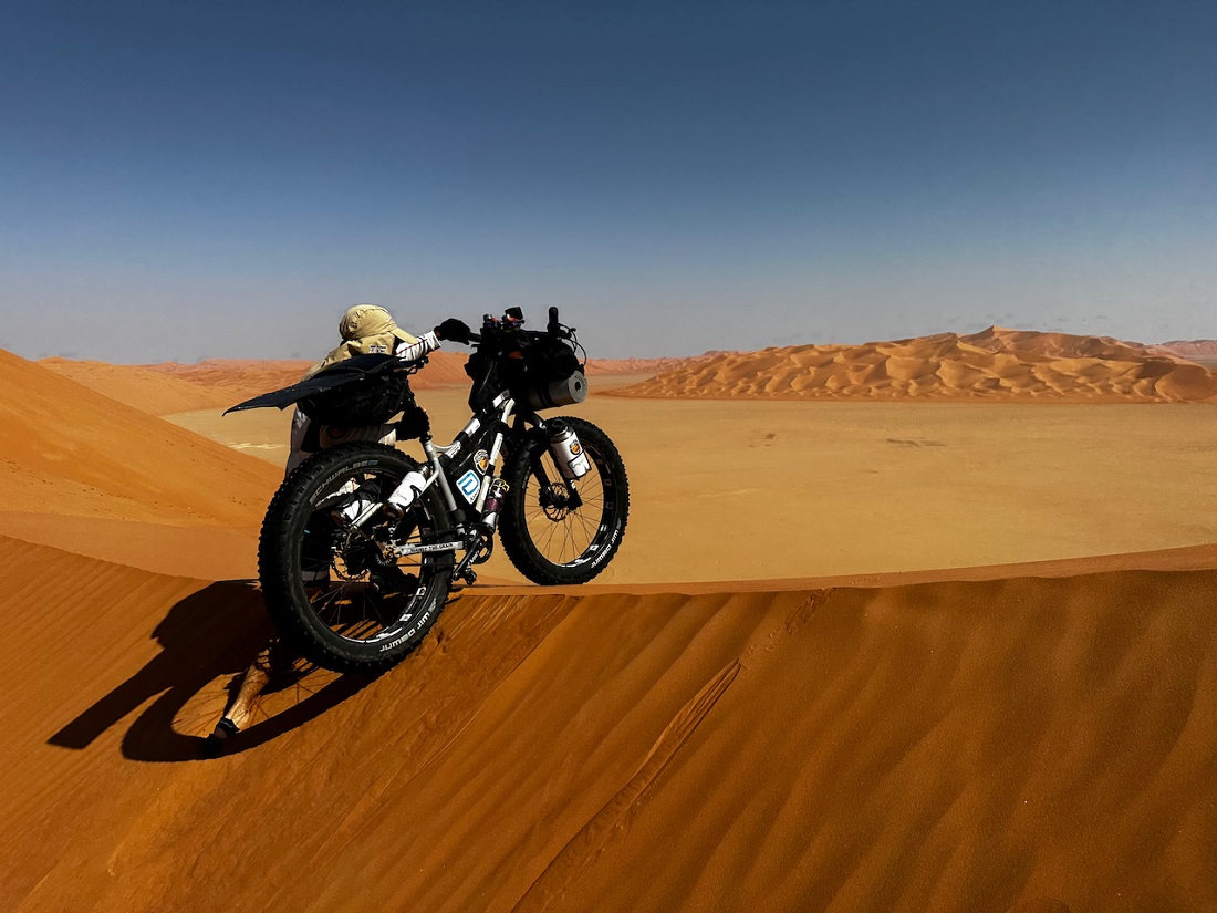 un homme pousse un vélo lourdement chargé sur une haute dune de sable 