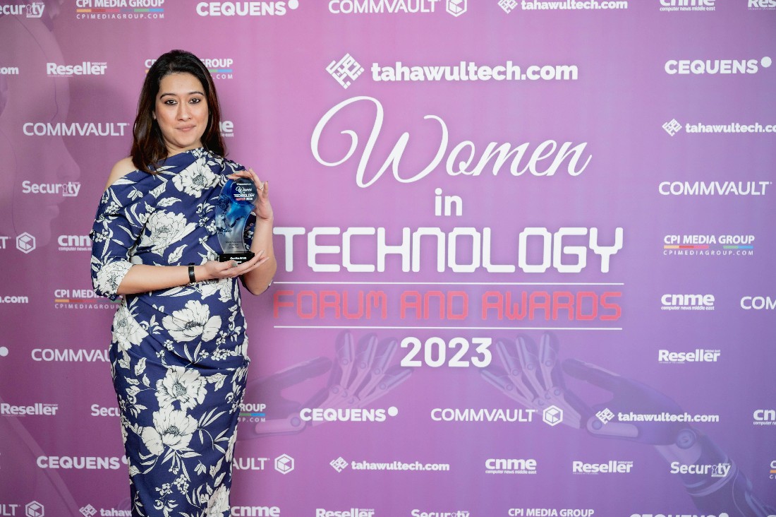 une femme vêtue d'une robe bleue à motif fleuri blanc tient un trophée devant un fond de présentation