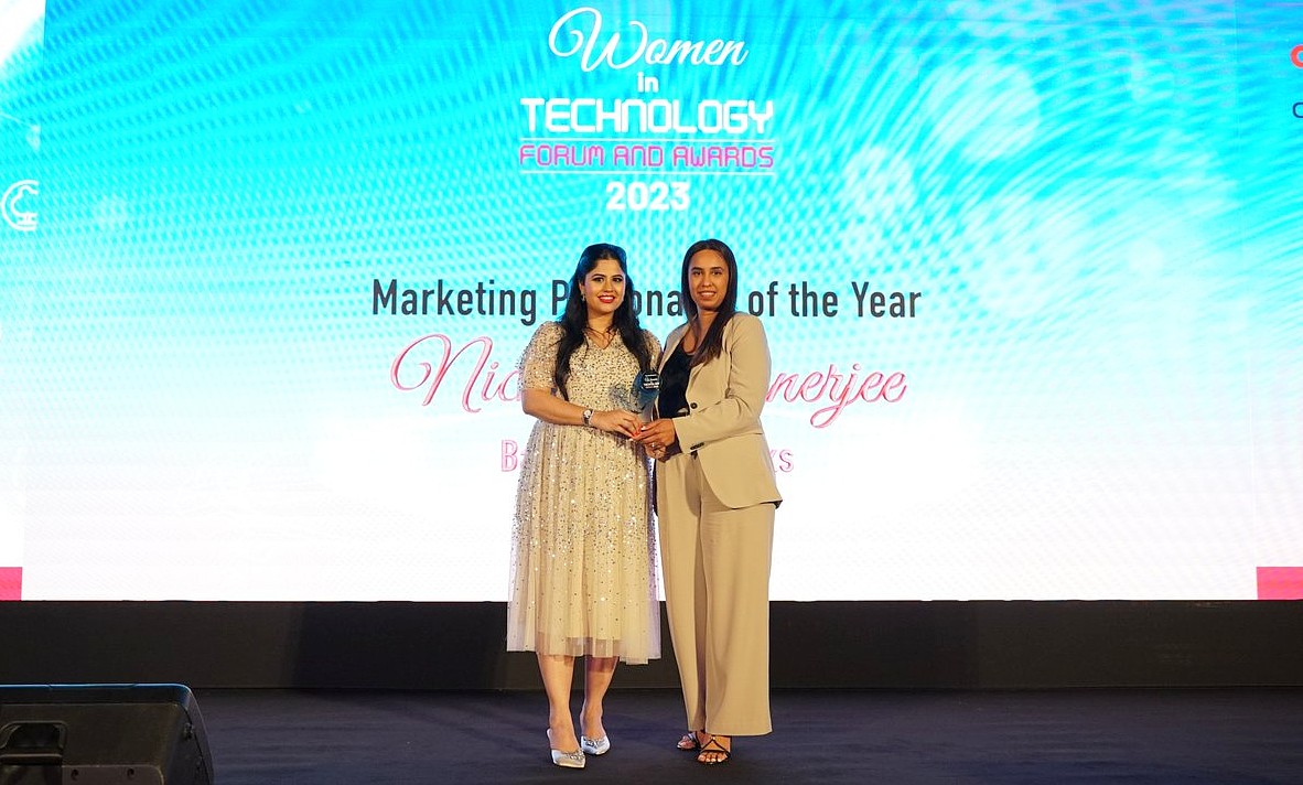 A trophy is presented to a woman standing on the right by a woman standing on the left of a presentation stage. 