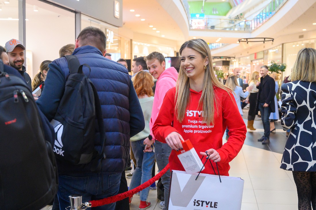 une femme portant un t-shirt rouge est vue en train de sourire à un client lors d'un événement d'ouverture d’un magasin de haute technologie