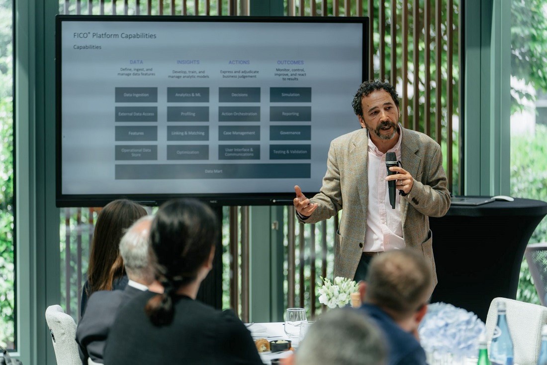 a man speaks into a mic as he presents to a small audience at a table