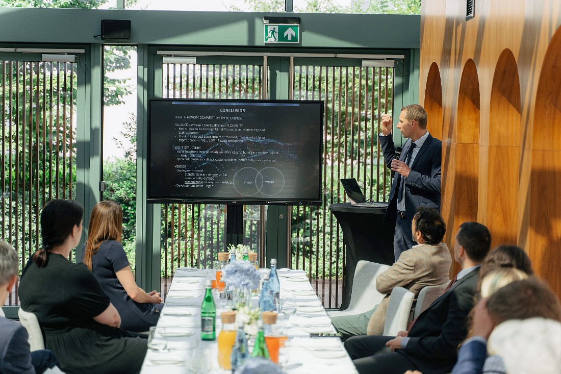 a man gestures to the presentation screen as he talks in front of a group sitting at a table. 