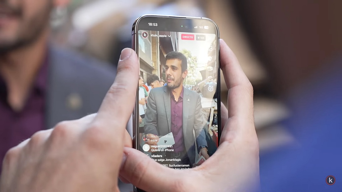 a magician is seen via a mobile phone performing a trick with a playing card