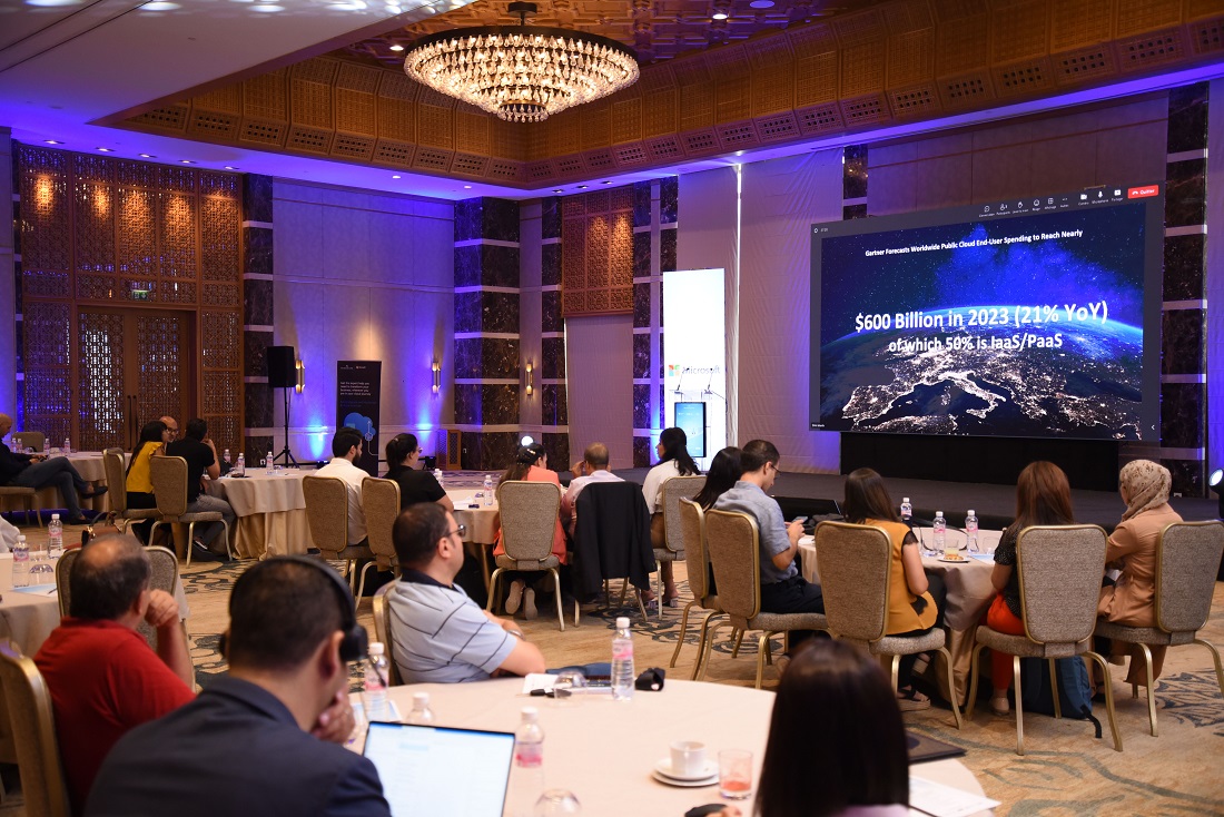 in a darkened room with some blue lighting, event attendees clustered around tables watch a presentation on a digital screen