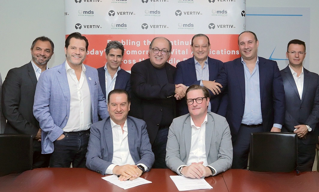 seven men stand in a group photo with two men in the foreground sitting at a table to sign an agreement