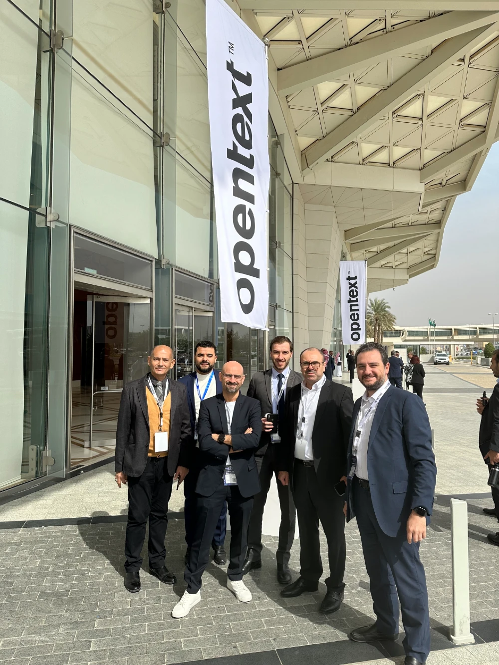 six men in suits are seen outside a conference venue with a banner spelling out the word, OpenText