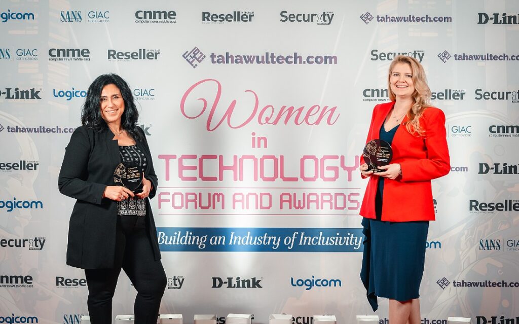 two women are seen holding trophies against a backdrop which spells out Women in Technology forum and awards