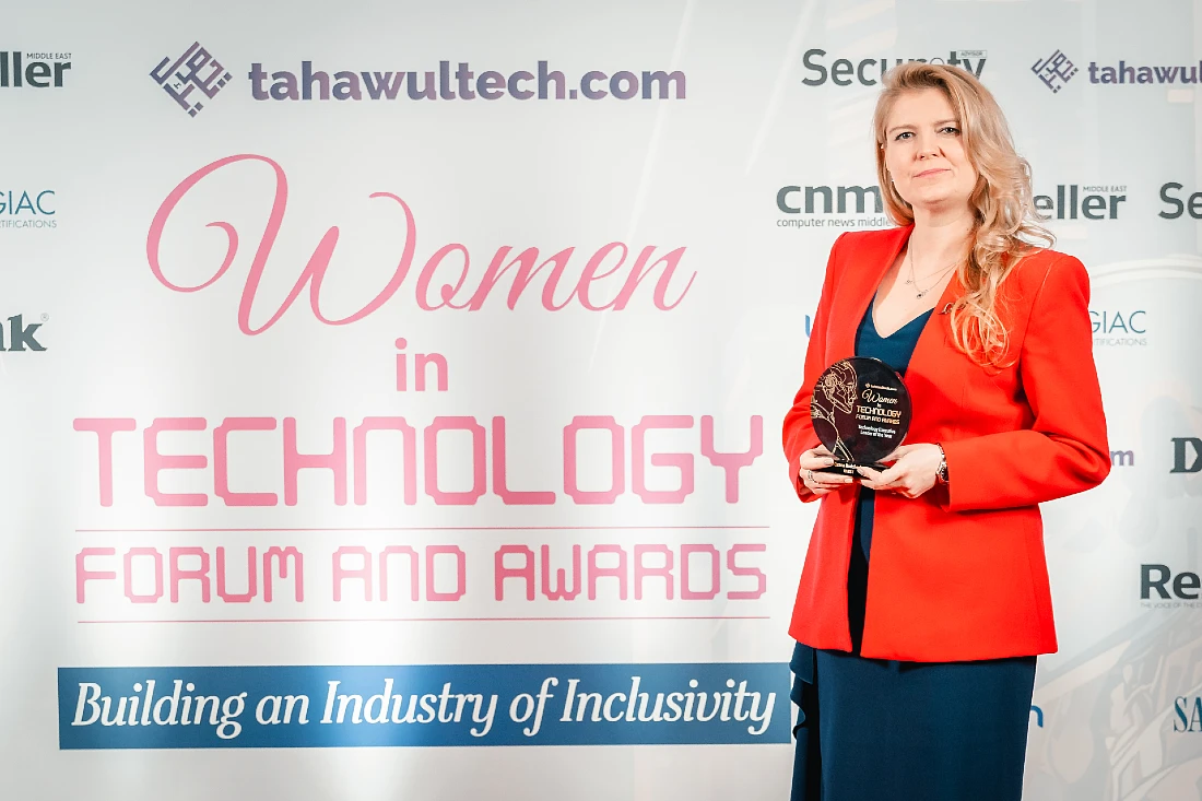 a woman in a red jacket holds a trophy in front of a screen with the words Women in Technology Forum and awards prominent