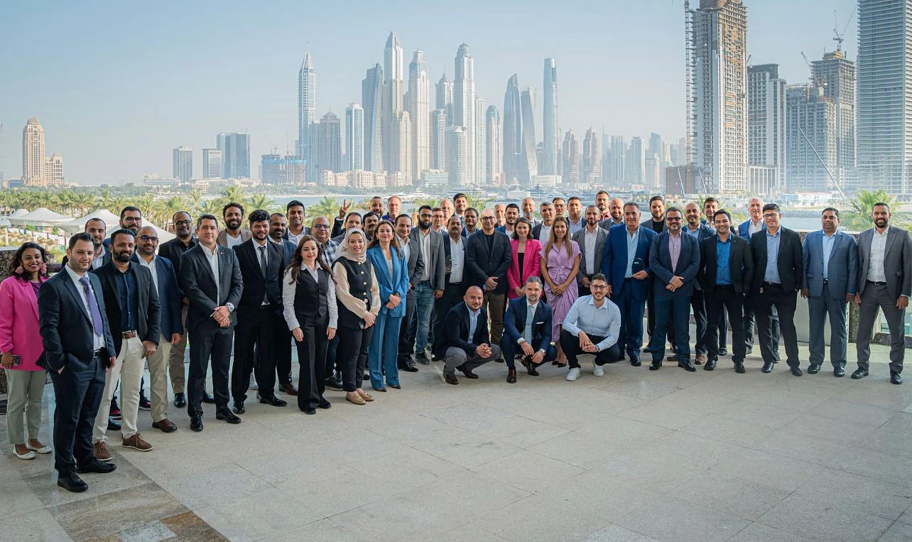 a large group of people gathered outside in a group photograph, with a city skyline seen in the background