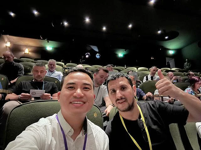 Two men are seen in a selfie in an auditorium