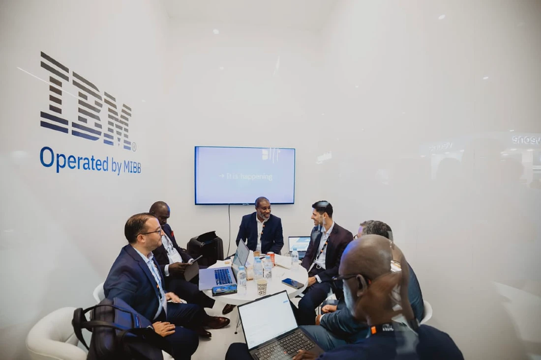 Six men in suits are seen working at a table with an IBM operated by MIBB logo on the wall behind them. 