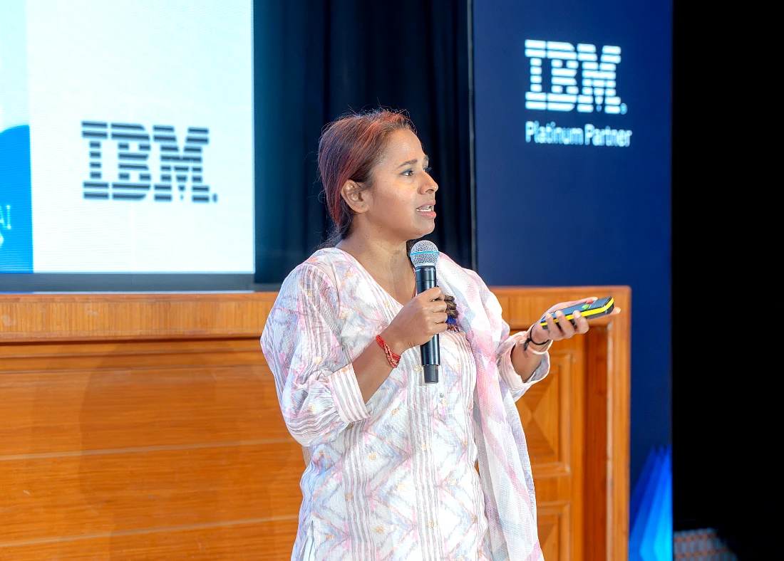 a woman speaks into a mic in front of a presentation screen with the IBM logo prominent
