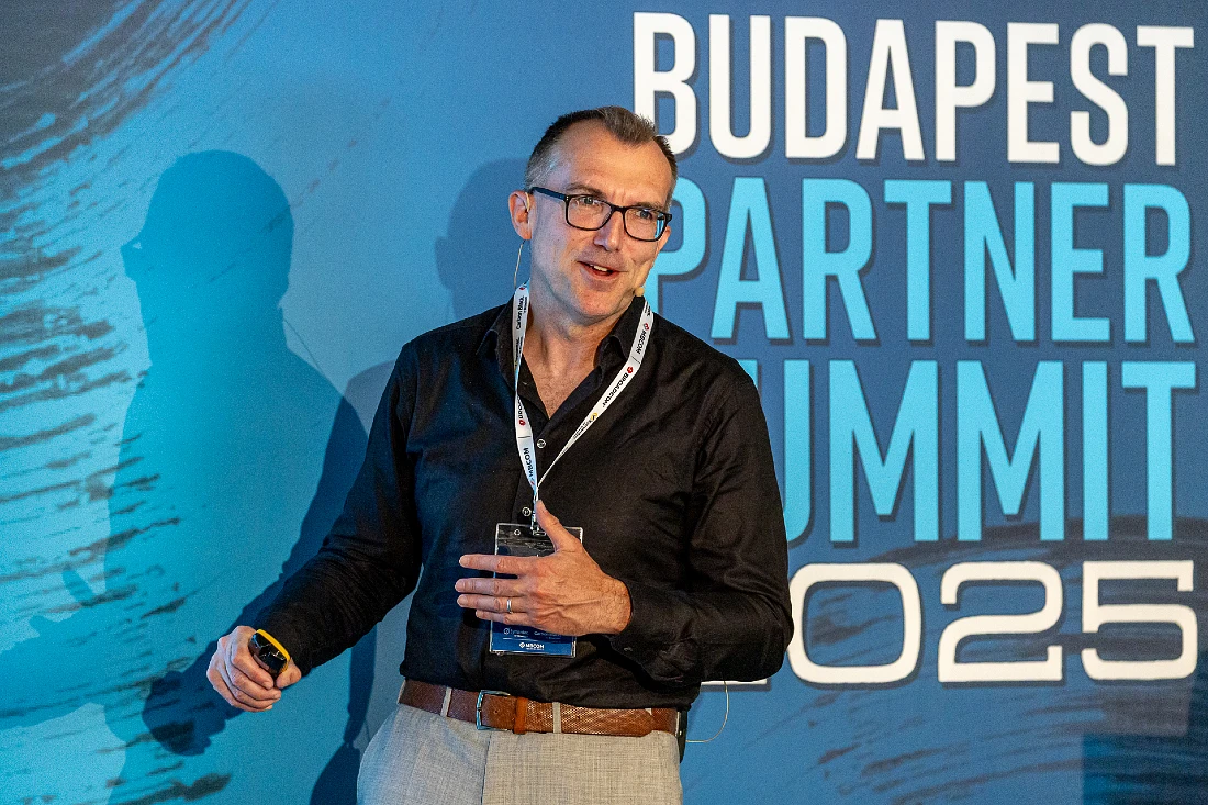 a man in a dark shirt is seen on stage presenting in front of a blue background with the words Budapest Partner Summit 2025 prominent behind him
