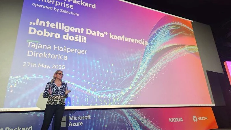 a woman presents a welcome speech to an audience while standing in front of a large digital screen