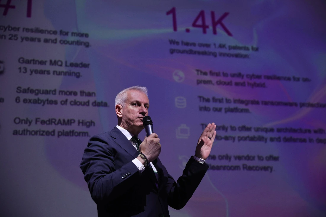 a man in a suit is seen speaking into a mic in front of a digital presentation screen
