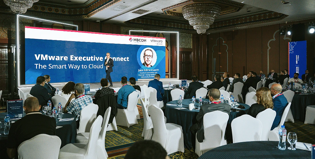 a man is presenting to an audience at a hi-tech event with people seen in the foreground watching the presentation