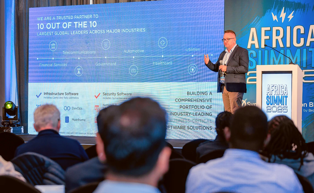 A man presents in front of a digital screen to an audience at an IT event