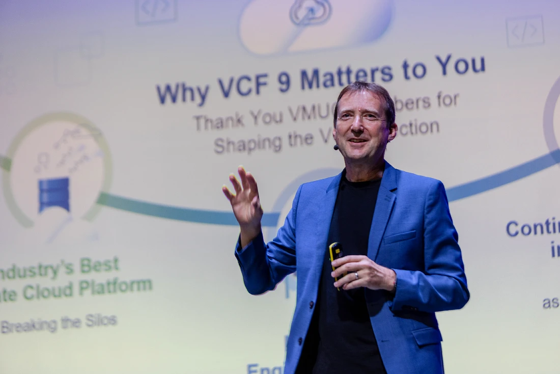 a man in a suit gestures as he presents in front of a digital screen at a hi-tech event 