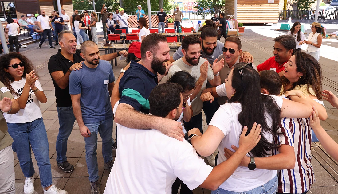 A group of people outdoors in casual clothes entwine their arms and form a circle, clapping and smiling at each other 