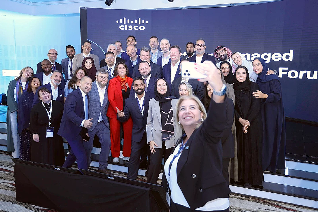 A group of colleagues are seen on a stage at a hi-tech event with one person in front taking a selfie