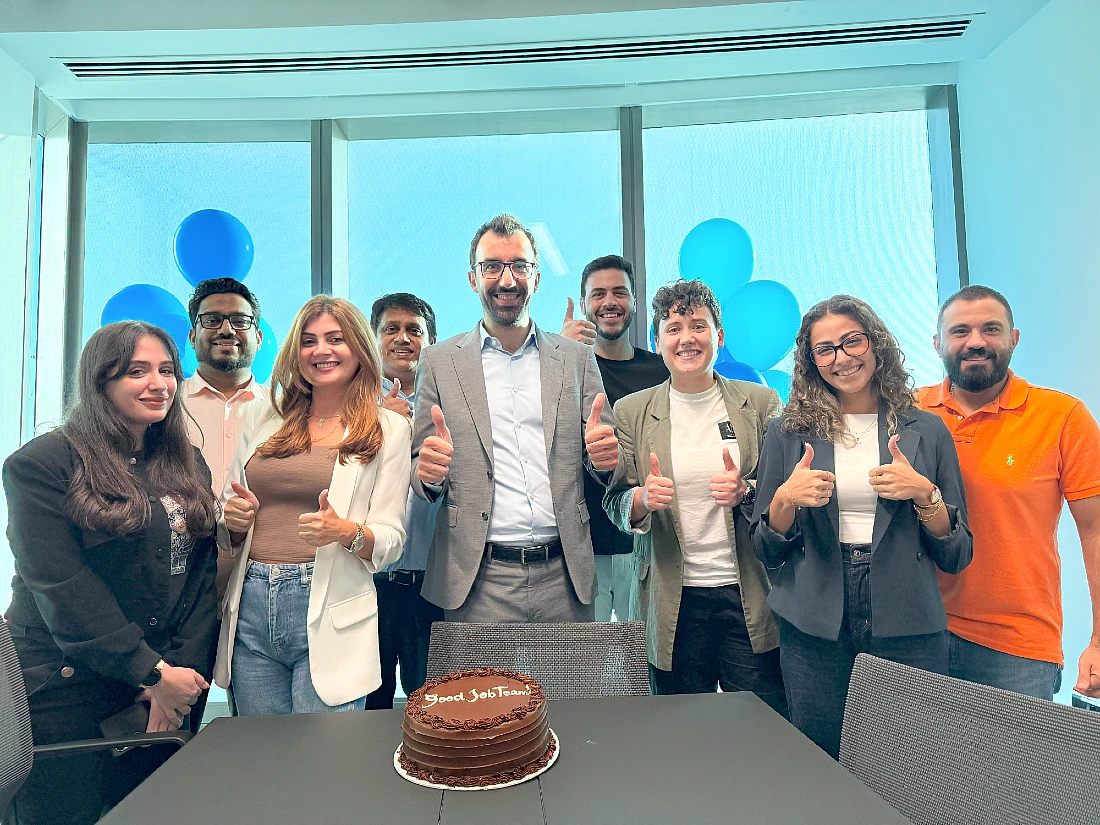 a group of colleagues in an office are seen smiling, with a cake in front of them on a table with the words Good Job Team spelt out in icing on the cake