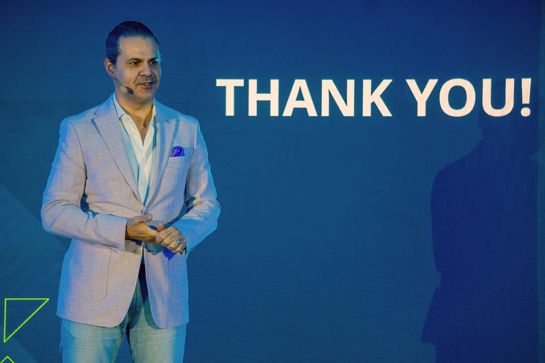 A man in a suit presents to an audience at a hi tech event with the words Thank You projected onto the background