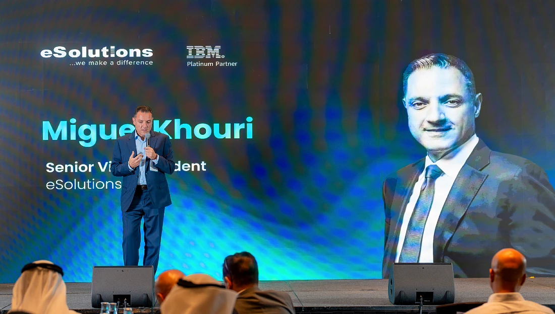 A man in a suit talks to an audience at a hi tech event with an image of himself projected onto the backdrop behind him
