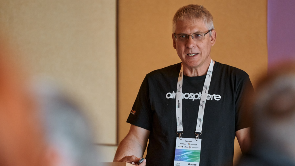 a man in a black t-shirt presents to an audience - he is wearing a lanyard around his neck.