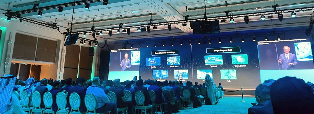 General view of an auditorium with many people seated in the foreground and a presenter talking on stage 