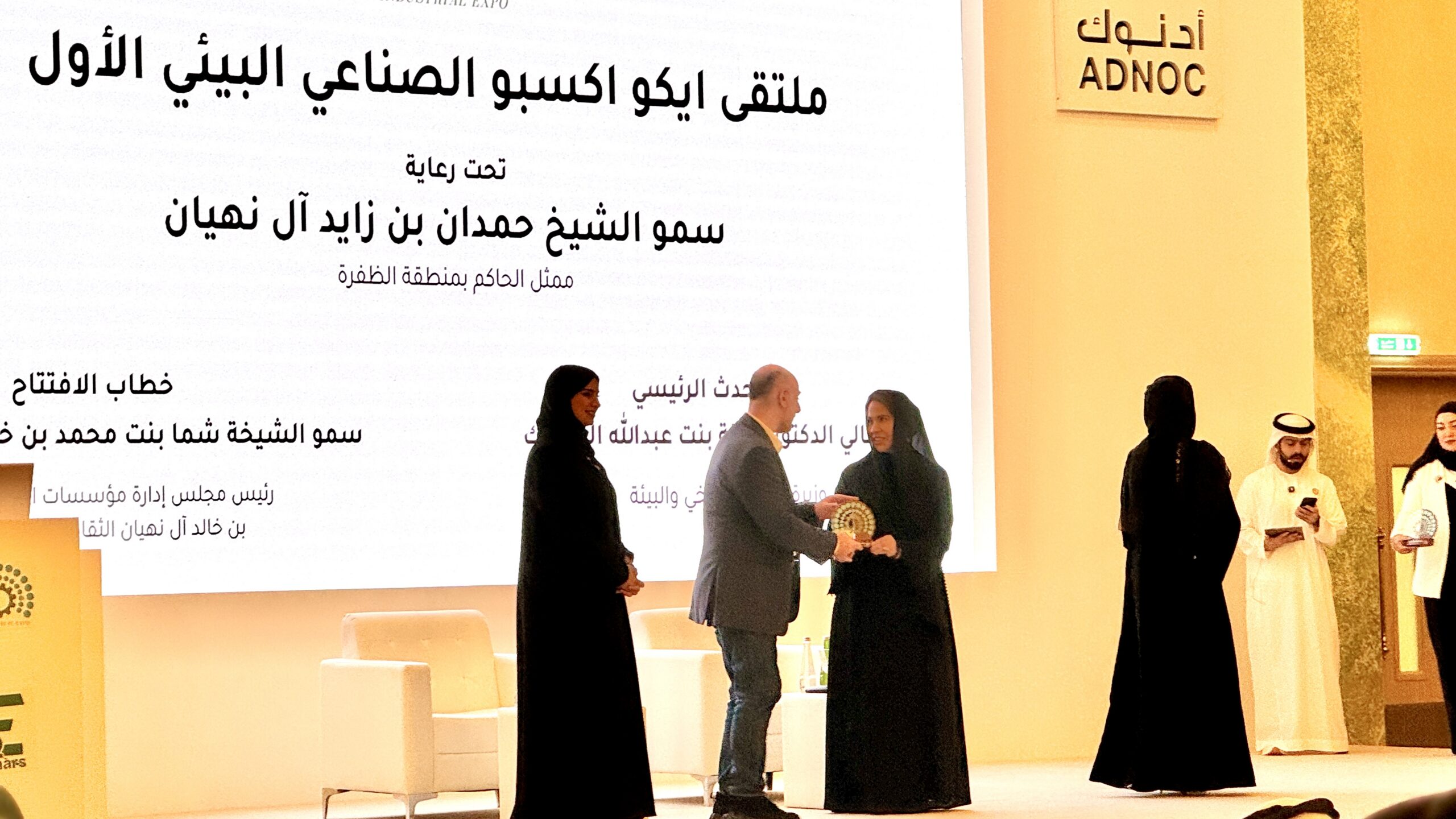 Six people are seen on a presentation stage as a plaque is presented to a man in a grey suit 