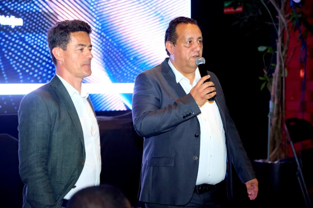 Two men in suits are seen with one of them talking into a mic at a hi tech event