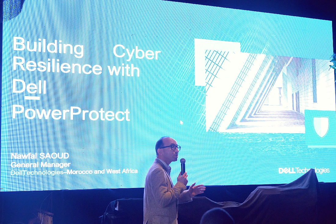 a man speaks into a mic in front of a digital presentation screen with a blue image on it