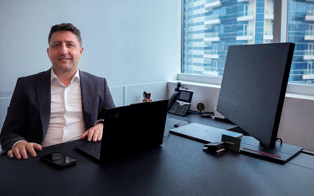A man in a suit sits at a desk with a computer nearby. He is looking directly at the camera.