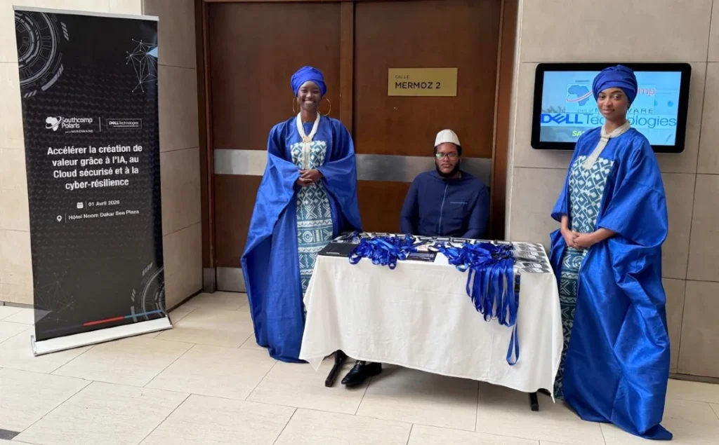 Deux femmes en robes bleu vif se tiennent à côté d'une table d'inscription à un événement où un homme est assis à la table.