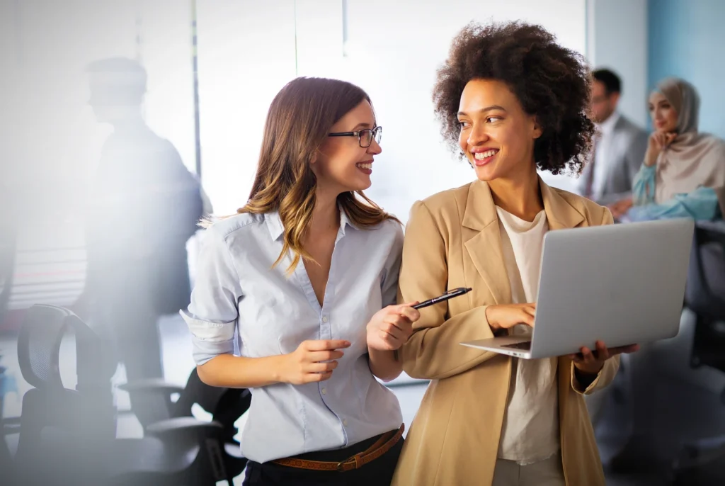 two women are talking to each other and smiling in a office setting. One of them holds a laptop.