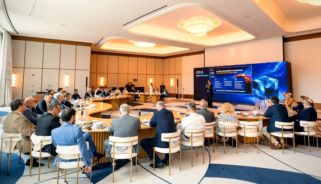 a roomful of people are seen gathered around a large, curved, irregular-shaped table at a hi tech event