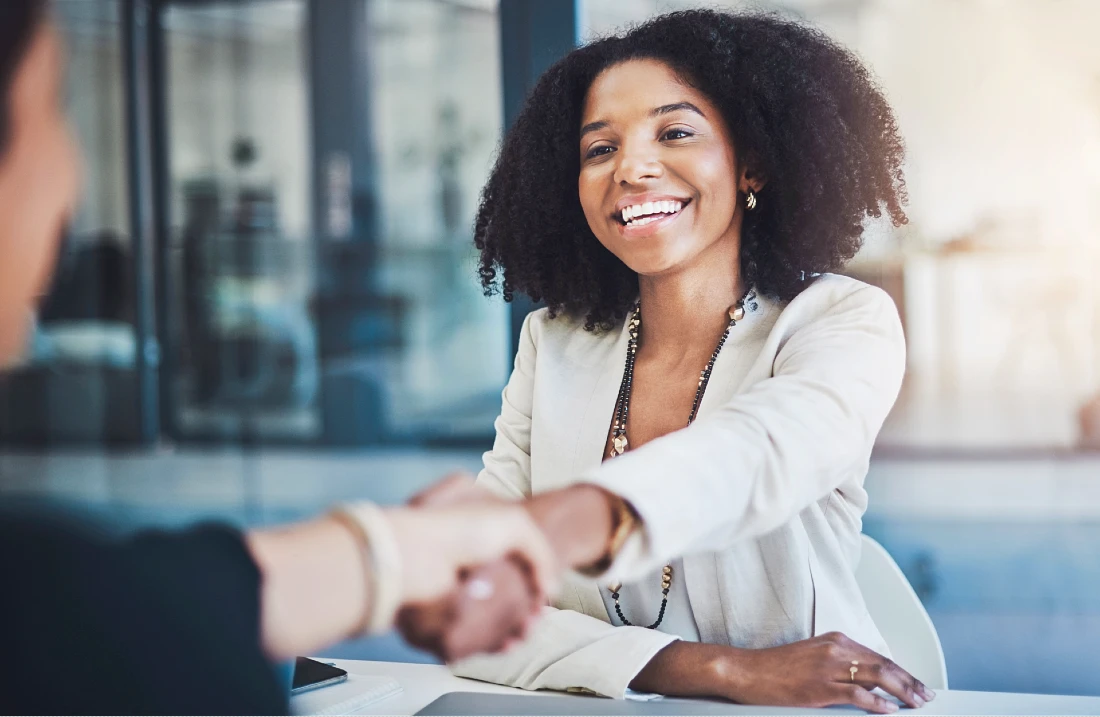 a woman in a business suit is seen shaking hands with a person on the left of the screen, who is out of focus.