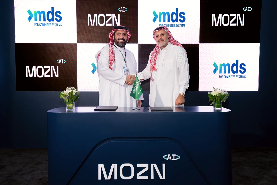 Two men are seen shaking hands in front of a logo covered screen